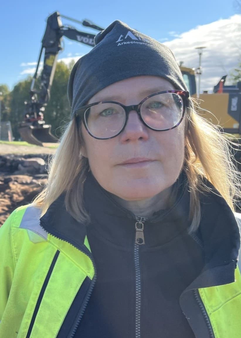 Annika Nordström står vid en byggarbetsplats där en grävmaskin arbetar med markberedning. Soligt väder och grönskande träd i bakgrunden.