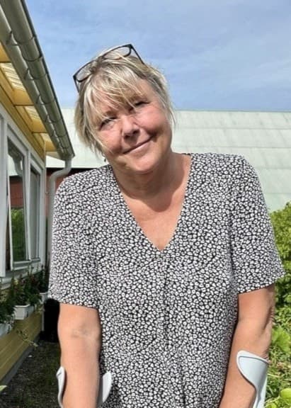 Åsa Enarsson står utomhus med kryckor vid en gul husfasad. Hon bär en blommig klänning och omges av växter, inklusive en solros i förgrunden. Högerbenet är förlamat efter borrelia.