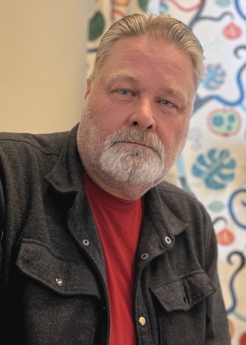 Christer Lindh från Venjan, Mora, sitter vid ett bord i ett rum med färgglada gardiner och ett fönster i bakgrunden. Han har en mörk skjorta över en röd tröja.