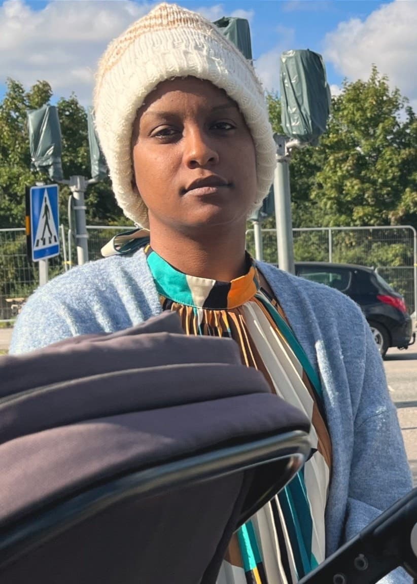 Ariam sitter med sin dotter i barnvagn vid ett övergångsställe i Gottsunda. I bakgrunden syns träd, höghus och en bil.