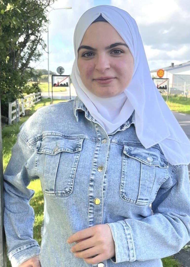 En bild som visar en förstörd mataffär efter en brand med taket kollapsat och brandrester runt byggnaden. Till höger står Rinad Khamis i en jeansjacka och vit hijab.