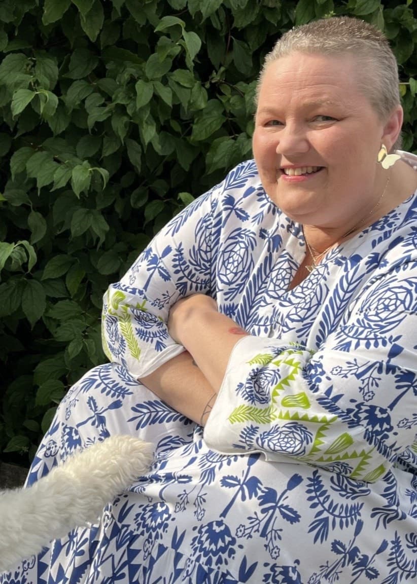 En person i en blåvit mönstrad klänning sitter utomhus vid en grön buske och interagerar med en vit lockig hund som står framför dem.
