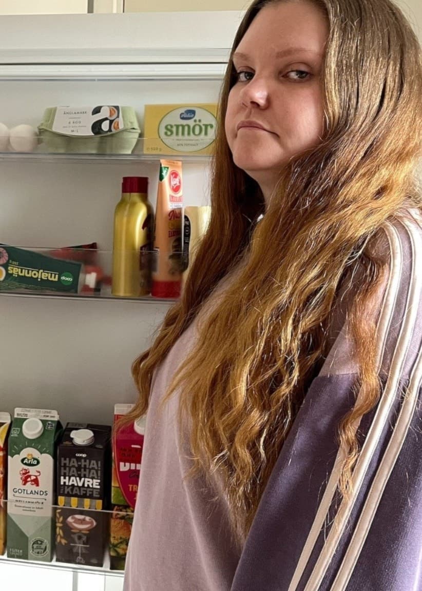 En kvinna står framför ett öppet kylskåp med varierande livsmedel, inklusive mejeriprodukter och juicer. Hon har långt hår och bär en randig tröja. Kylskåpet innehåller flera varor med kort utgångsdatum.
