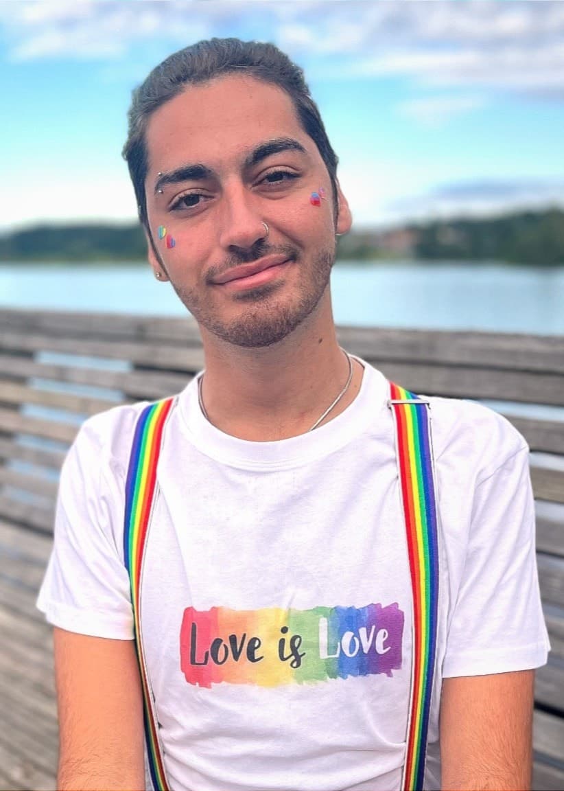 Anwar Ghoubari sitter på en träbrygga vid vattnet, klädd i en vit t-shirt med regnbågsfärger och hängslen. Himlen är blå med några moln i bakgrunden.