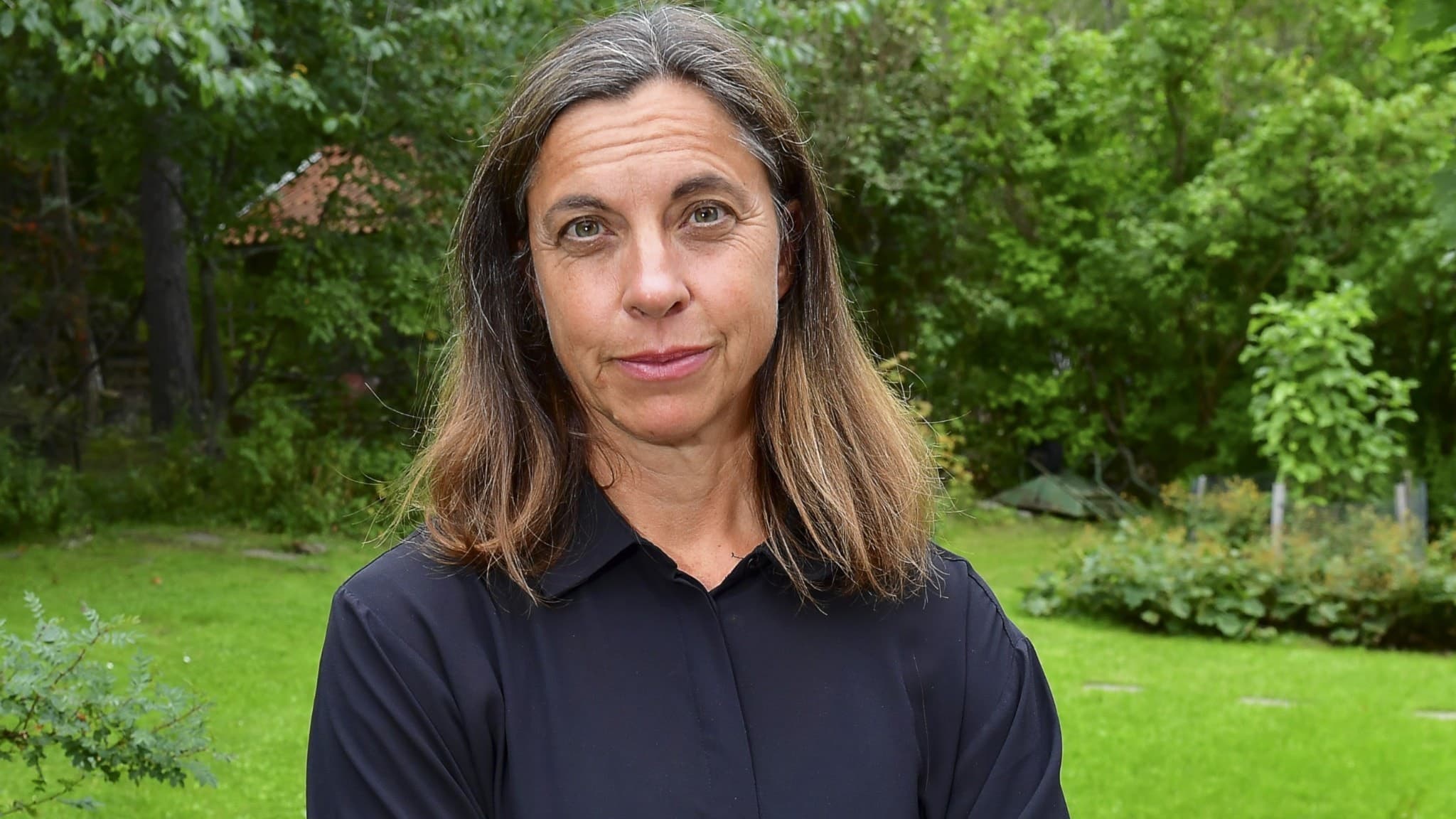 Hanna Thermaenius, barnpsykolog på Rädda Barnen, står med armarna i kors i en grönskande trädgård. Hon har på sig en mörk skjorta och omges av träd och växtlighet i bakgrunden.