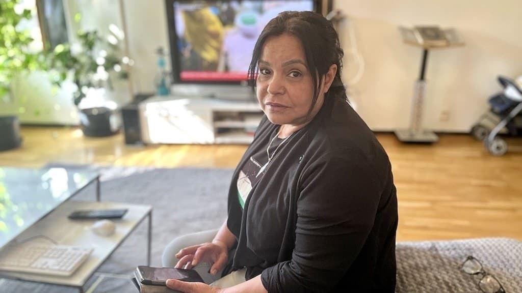 Jasmine Felestin sitter i ett vardagsrum med en smartphone i handen. Bakgrunden visar en tv som sänder nyheter och en ljus, modern inredning med växter och möbler.