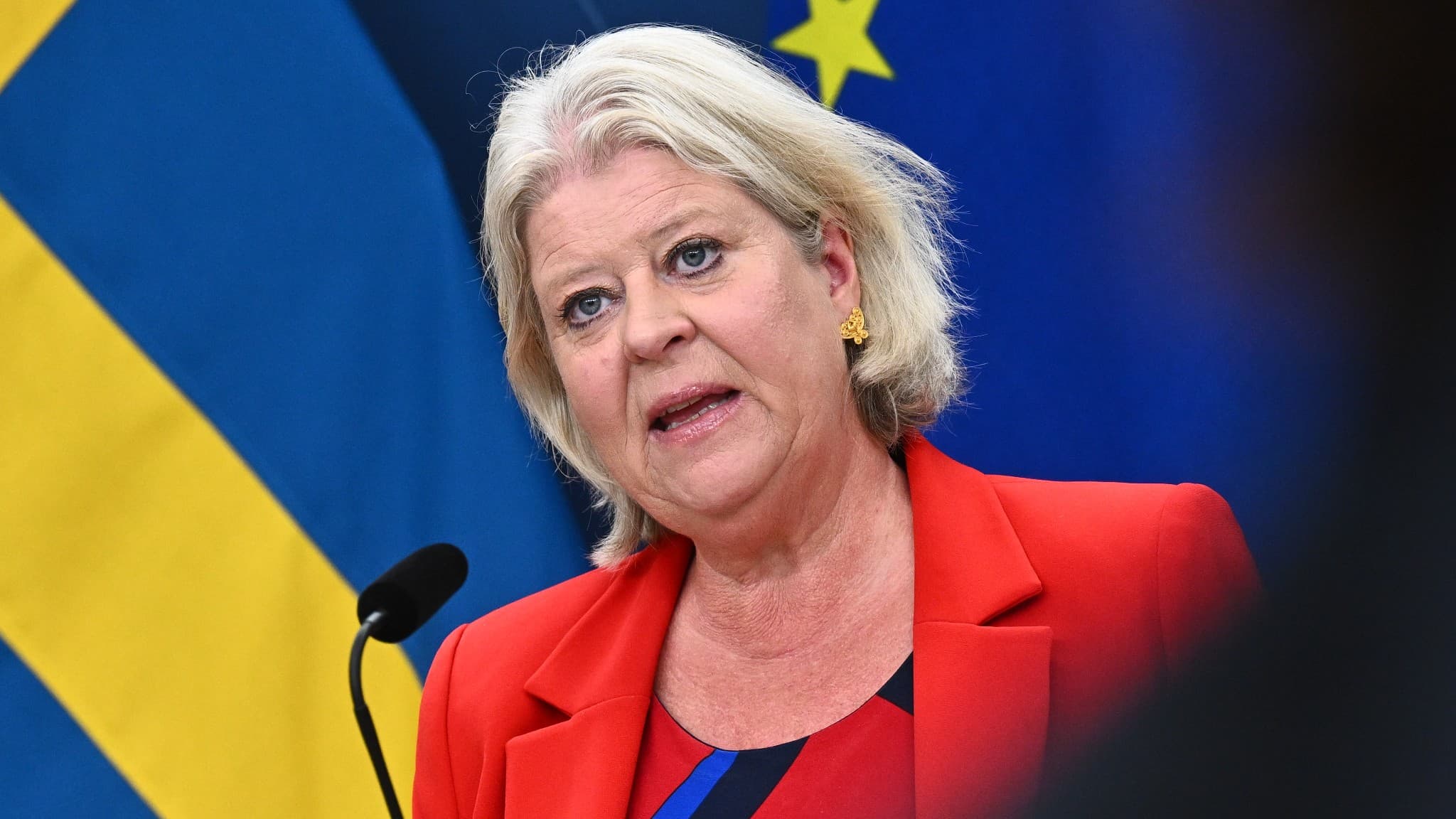 Socialtjänstminister Camilla Waltersson Grönvall (M) talar vid en pressträff. Hon bär en grön kavaj och står framför en svensk flagga i bakgrunden.