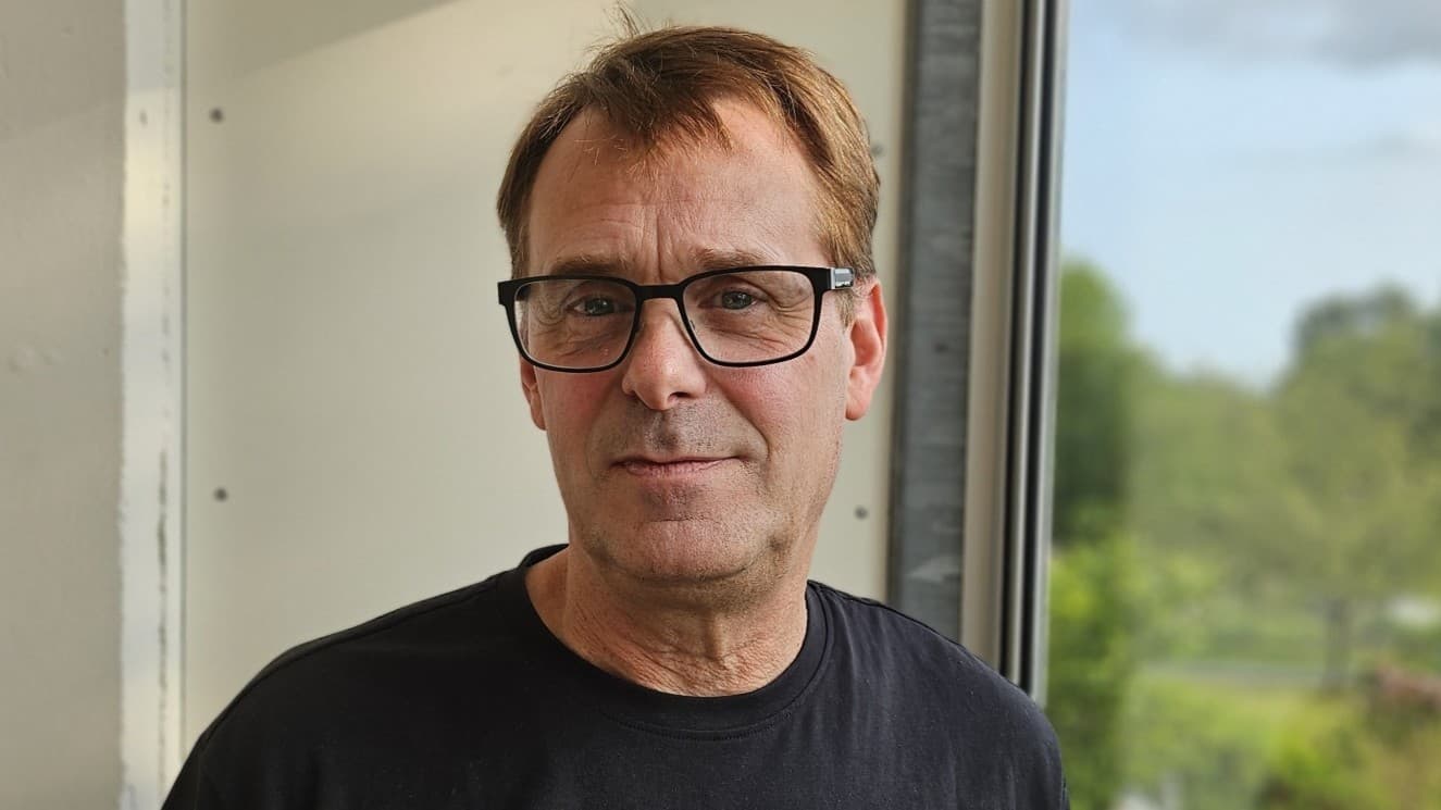 Peter Gustavsson står vid ett fönster i en svart tröja med en logotyp, bredvid en dammig industrimiljö där ljusstrålar tränger genom taket.