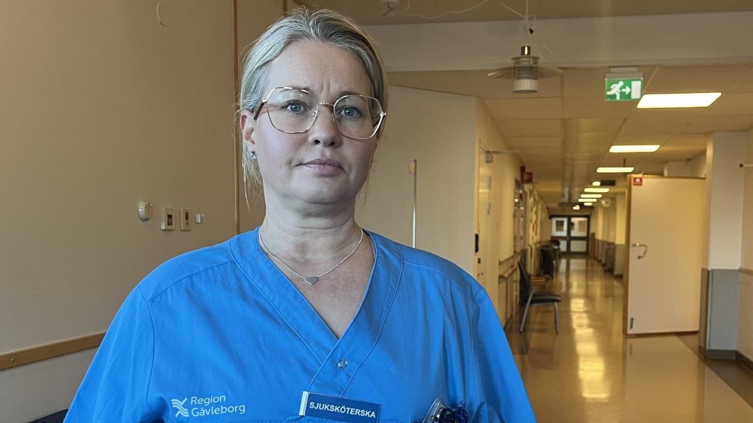 Sjuksköterskan Linda Gillberg på Familjeläkarjouren i Gävle står i en tom sjukhuskorridor. Hon bär blå arbetskläder och glasögon.