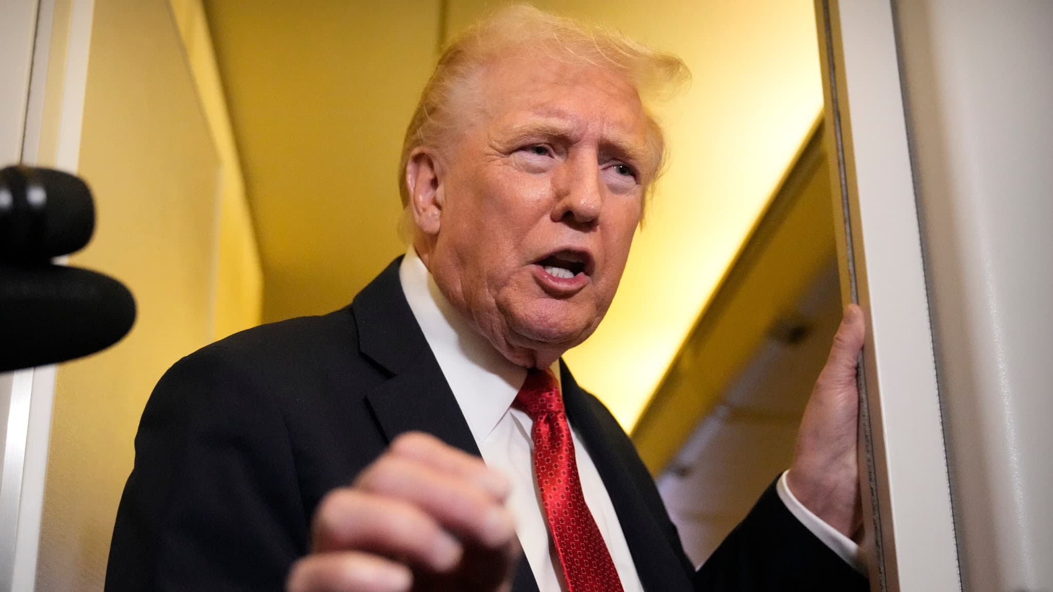 USA:s president Donald Trump iförd kostym och röd slips, talar till reportrar ombord på Air Force One, med mikrofoner synliga i förgrunden.
