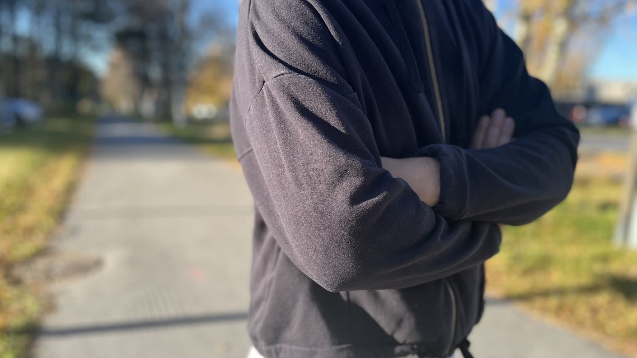 ”Kalle” står utomhus med korslagda armar, klädd i en mörk hoodie. Bakgrunden visar en gångväg med höstfärgade träd och gräsmatta i dagsljus.