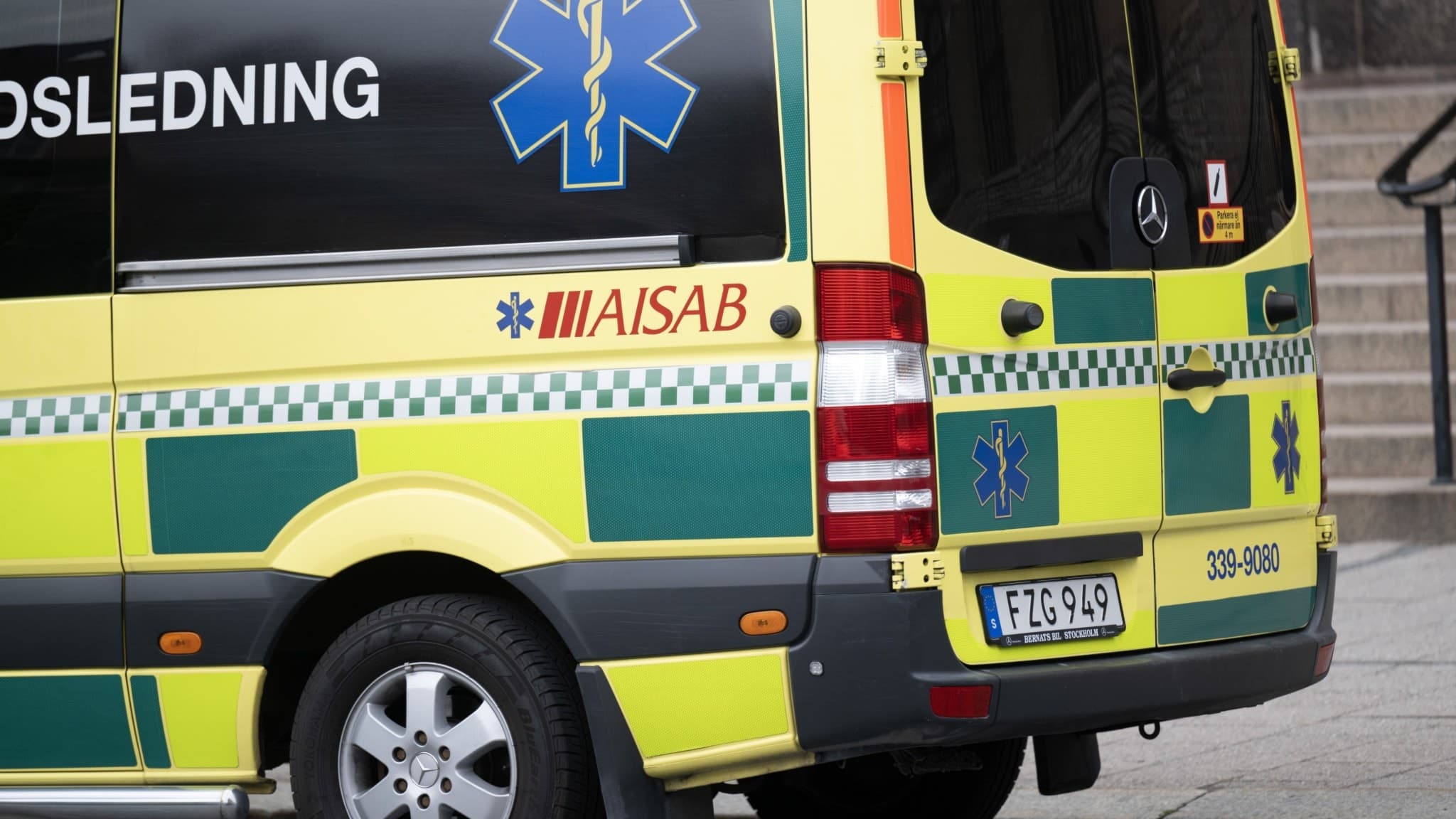 En gul och grön ambulans med texten 'SOS-ledning' och AISAB-logotypen syns parkerad vid en trottoar, med fokus på bakdelen av fordonet. Ambulansens blåa symbol syns tydligt.