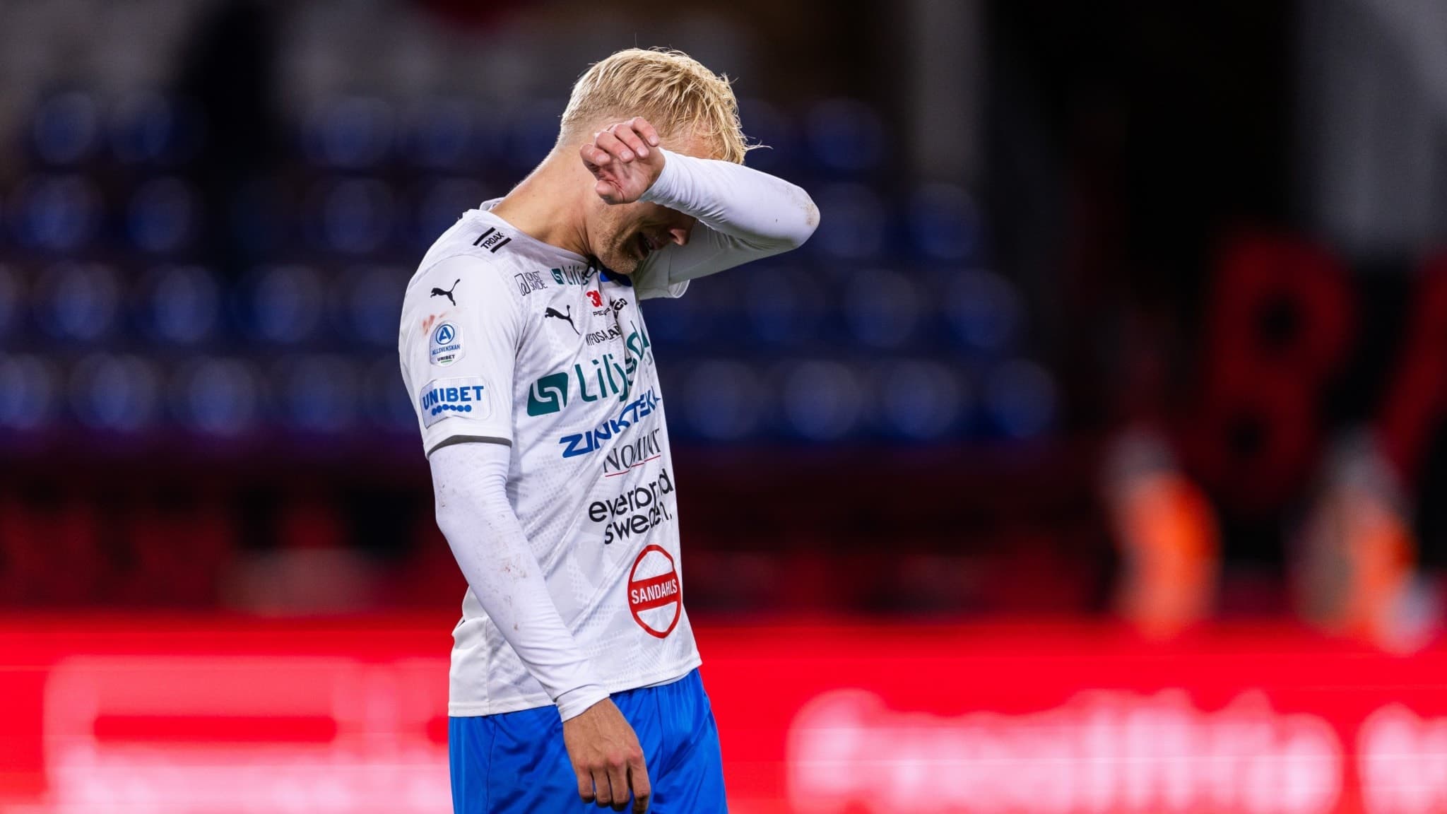 Fotbollsspelare i vit tröja och blå shorts står och deppar