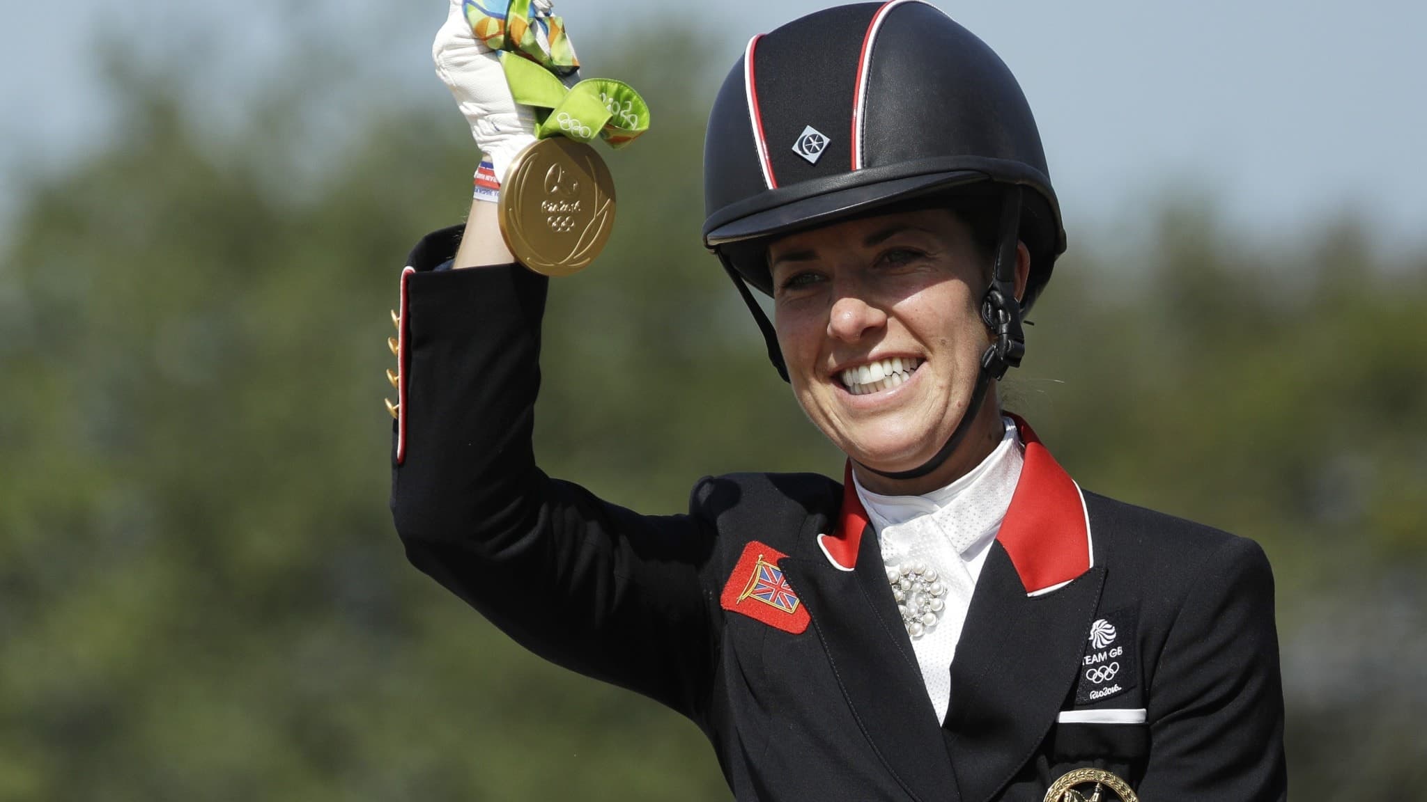 Charlotte Dujardin, iförd svart ridkavaj och hjälm, håller upp sin guldmedalj efter att ha vunnit individuell dressyr vid OS i Rio de Janeiro 2016. En grön medaljband syns tydligt.