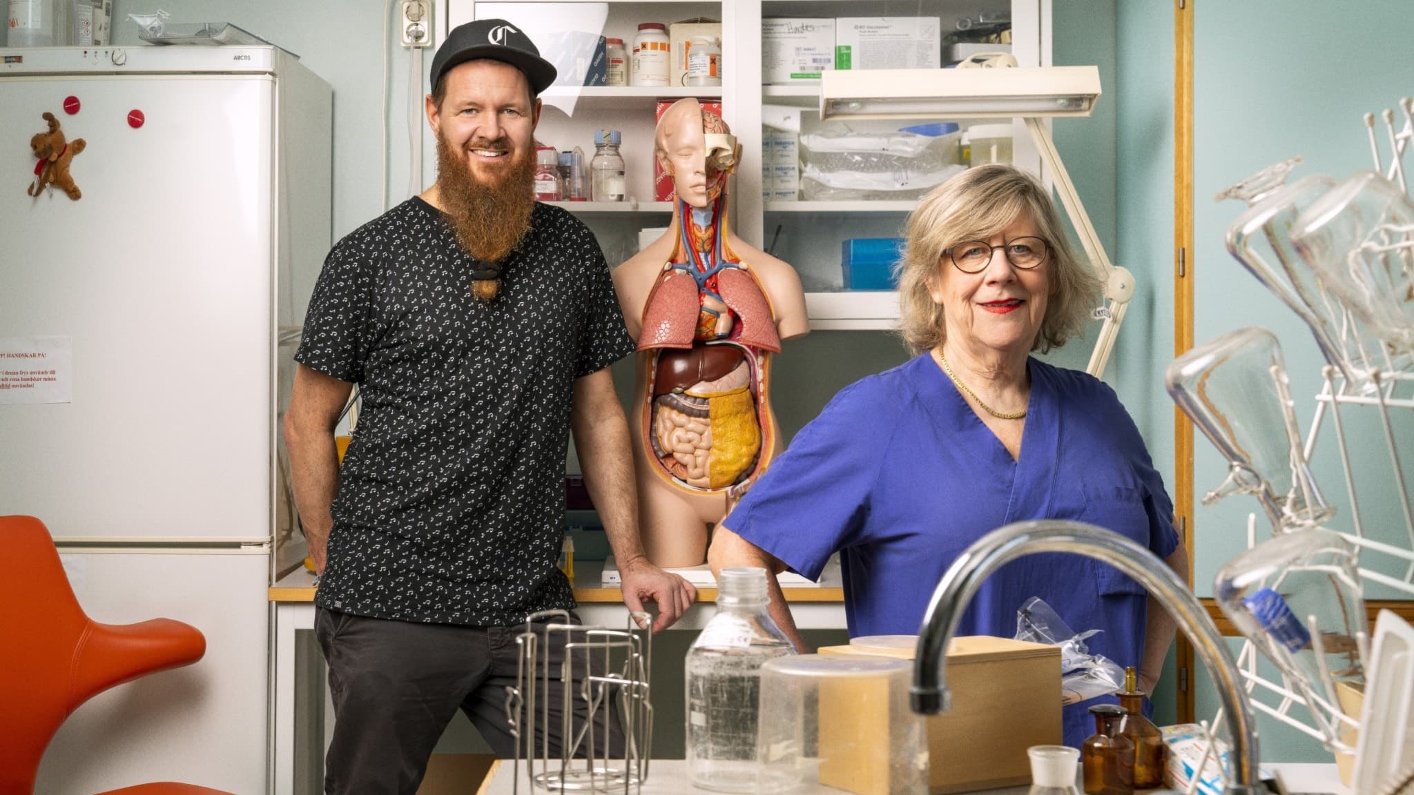 Christer Lundberg och Agnes Wold ståendes i ett laboratorium, Christer med skägg och keps till vänster i bild, till höger om honom en anatomisk modell över en människokropp. I förgrunden, till höger, står Agnes Wold med en blå läkarskjorta och glasögon.