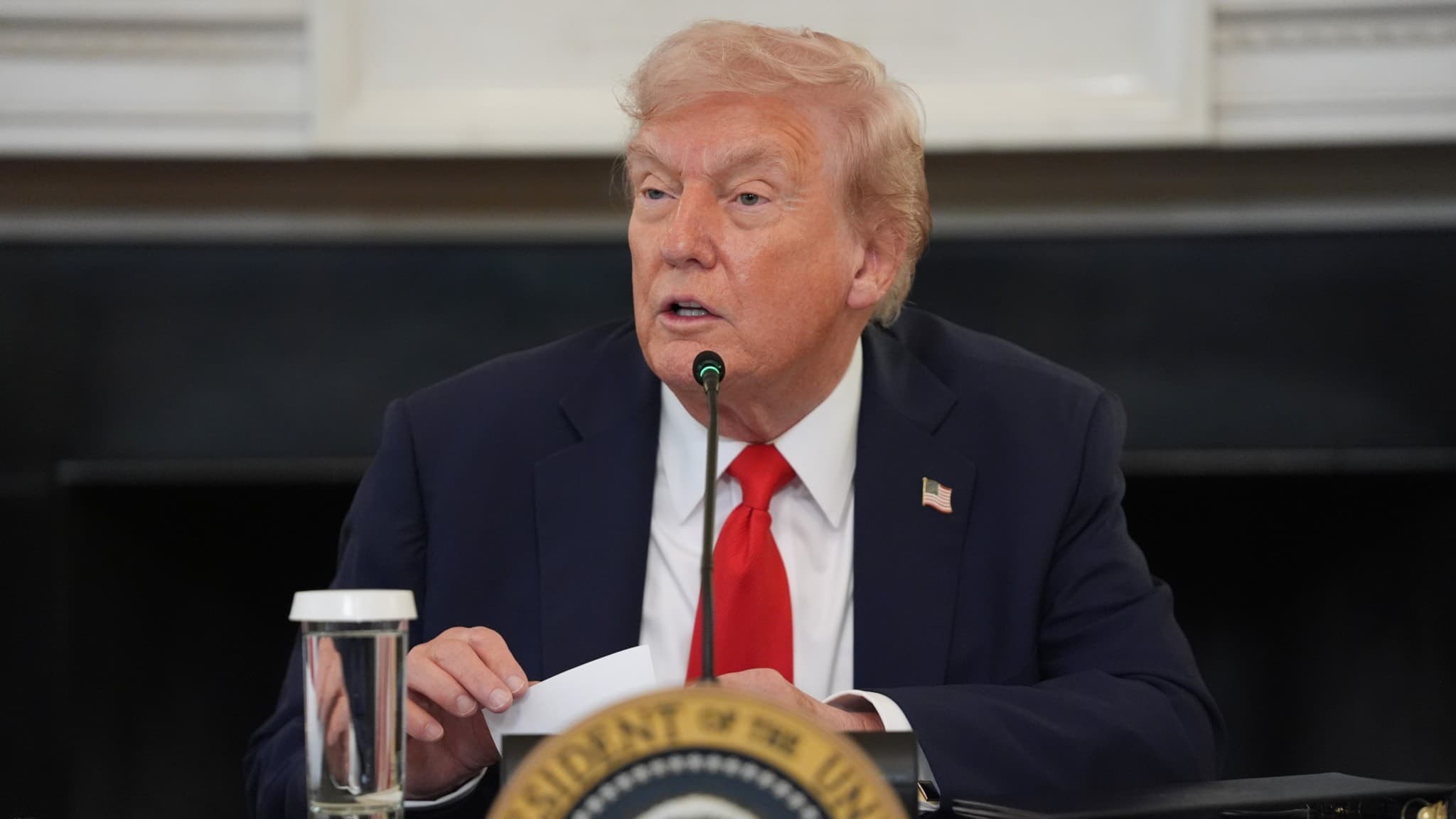 Donald Trump sitter vid ett bord i Vita husets State Dining Room, iklädd kostym och röd slips, med ett dokument i handen och ett glas vatten bredvid sig. En presidentemblem syns på bordet.