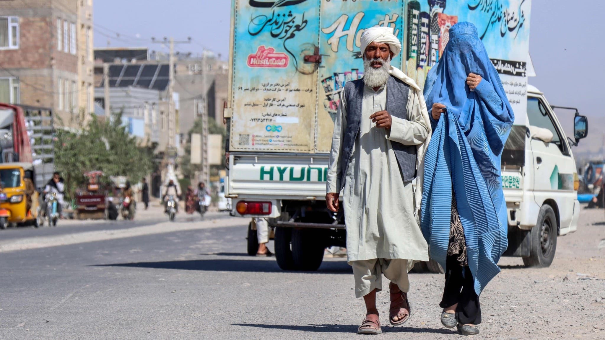 En afghansk man i traditionella kläder går bredvid en burkaklädd kvinna på en gata i utkanten av Herat. I bakgrunden syns en lastbil med färgglada reklamaffischer och stadsmiljö.