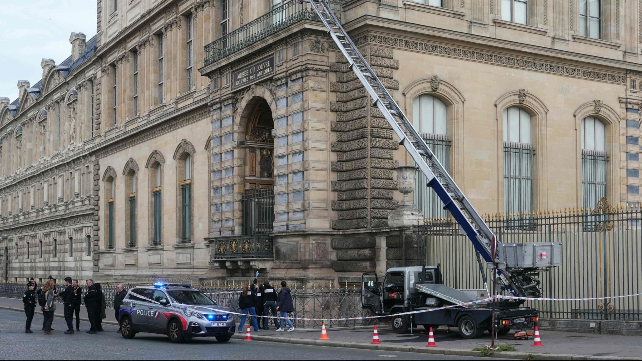 Poliser vid Louvren i Paris undersöker en möbelhiss som användes vid ett rån. Området är avspärrat med koner och avspärrningsband och flera tjänstebilar står parkerade vid platsen.
