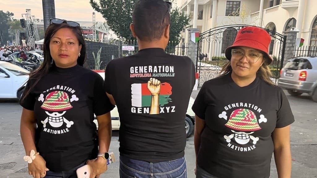 Tre personer klädda i svarta t-shirts med texten 'Generation Nacionalista' och en mexikansk flagga står utomhus vid en gata, med en byggnad och bilar i bakgrunden.