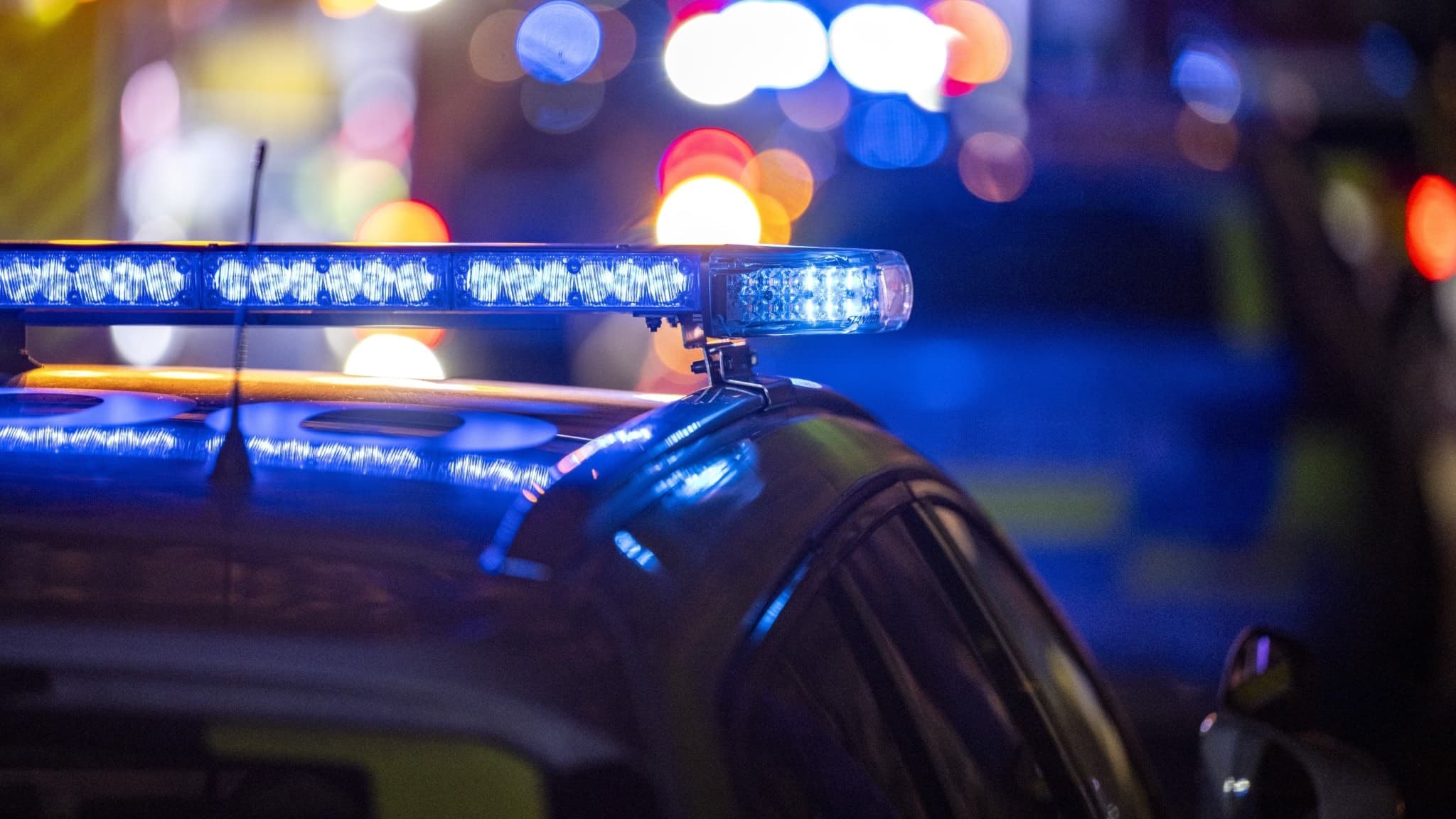 Polisbil med blåljus påslagna i mörker, mot en suddig bakgrund med fler ljus i olika färger. Bilden förmedlar en känsla av insats eller utryckning, relevant för brottsrapportering.